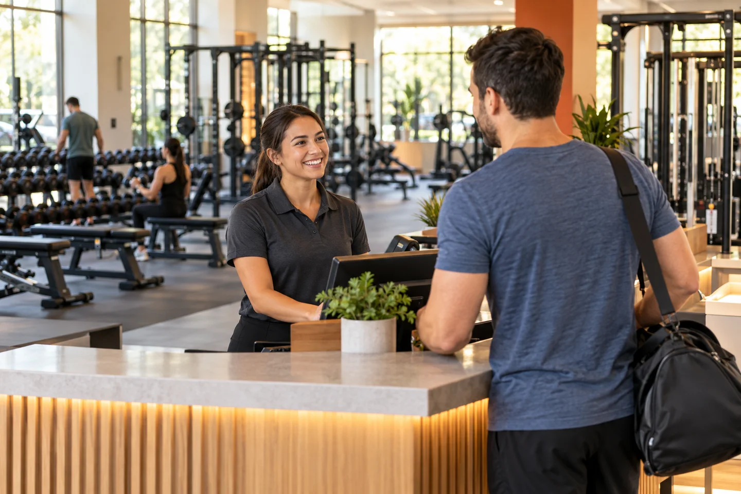 A gym staff member welcoming a member at reception with fitness equipment behind them